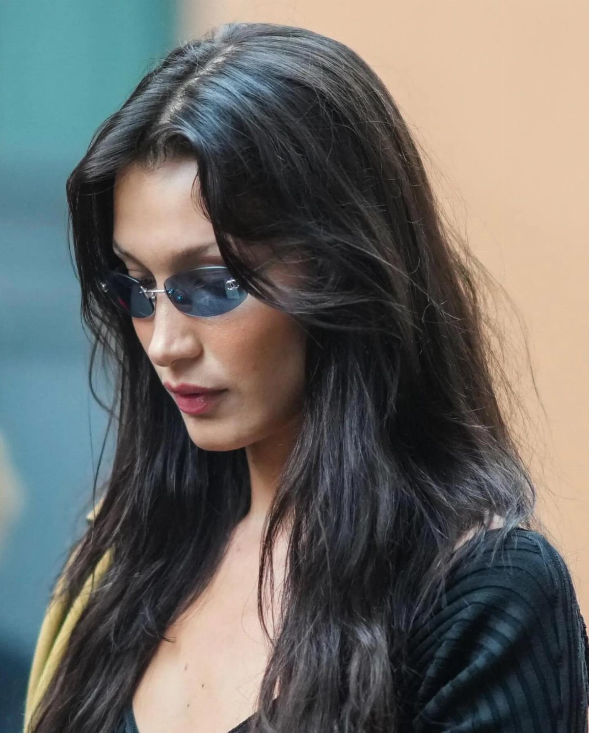 A woman with long dark hair wearing small oval tinted sunglasses and a black ribbed top.