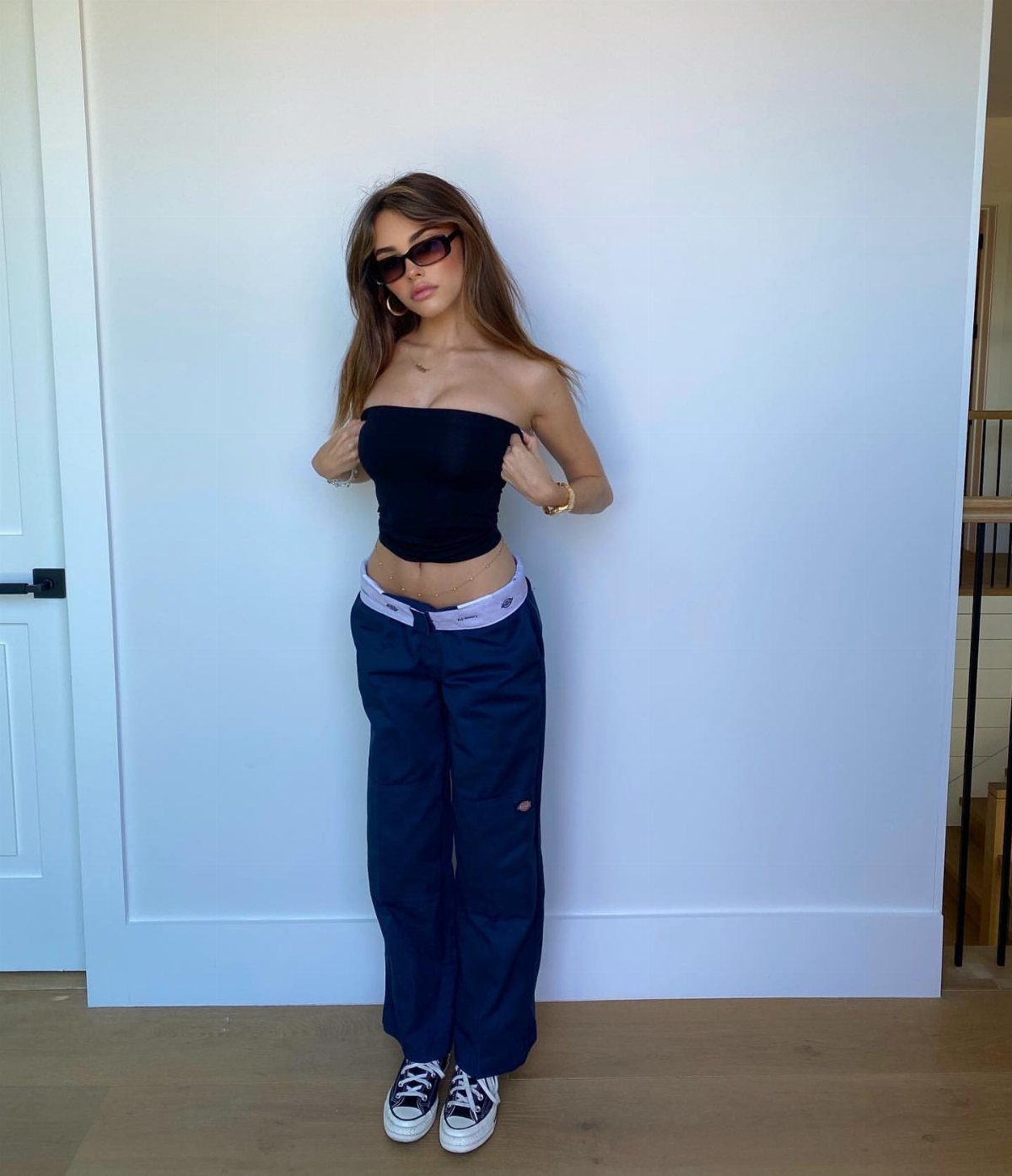 A woman wearing a black strapless tube top and navy blue cargo pants against a white wall.