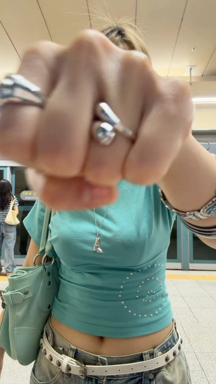 A person wearing a teal crop top, studded belt, and silver rings holding a fist forward.