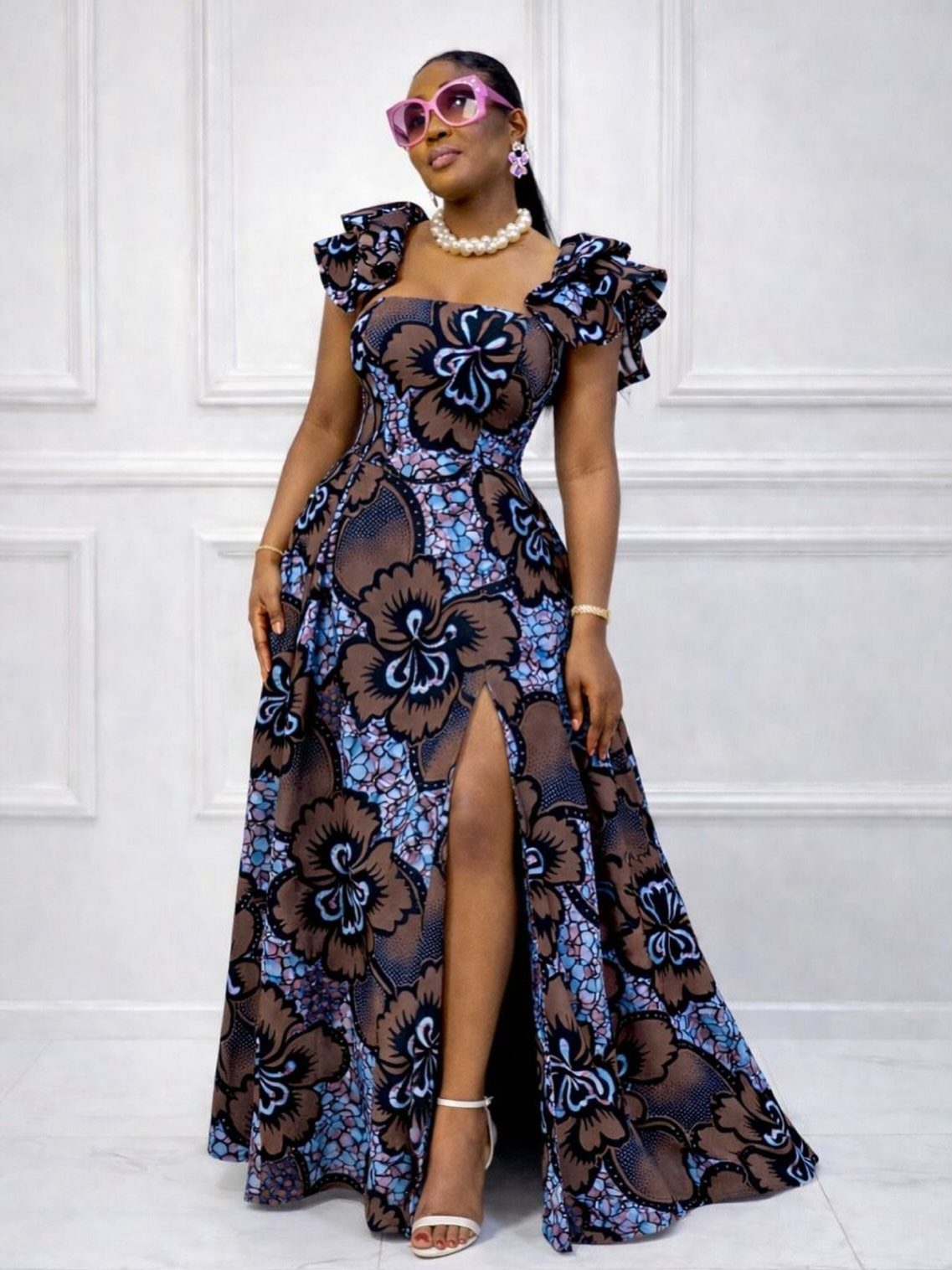 A woman wearing a floral A-line maxi dress with ruffles and a high slit posing against a white wall.