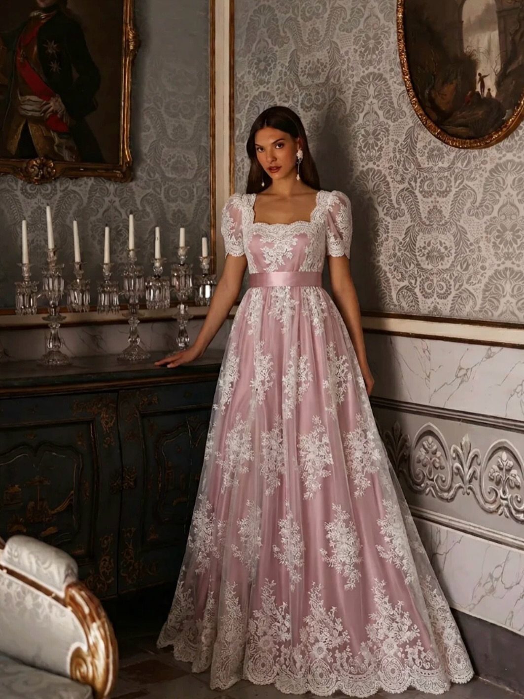 Woman wearing a pink lace empire style gown standing in an ornate room.