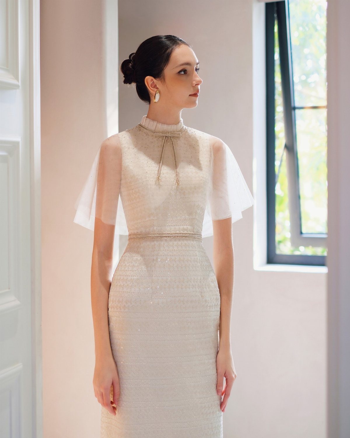 Woman in a shimmering cream high-neck dress with a sheer tulle capelet standing by a window.