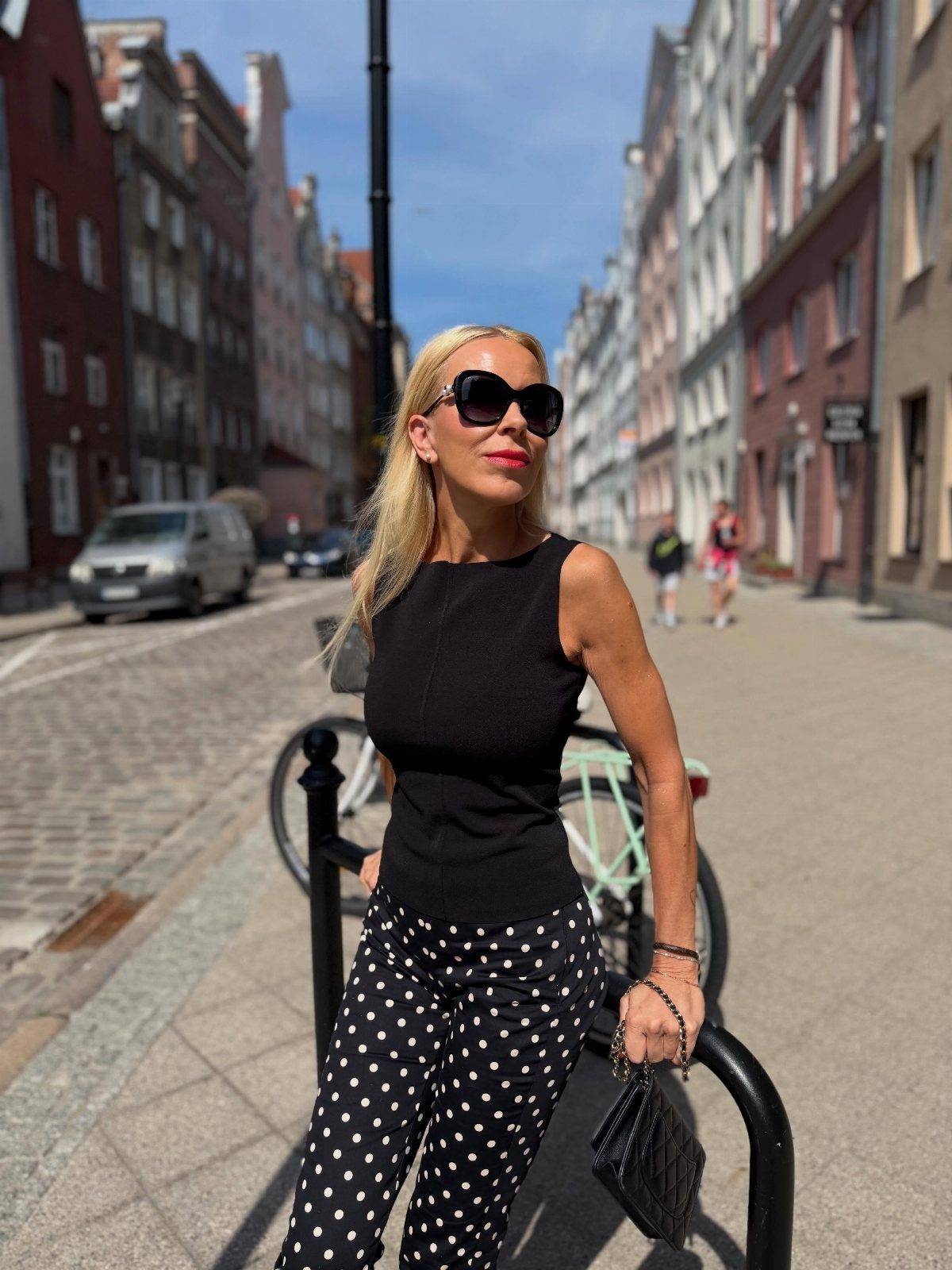 A blonde woman in a black sleeveless top and polka-dot pants posing on a European street.