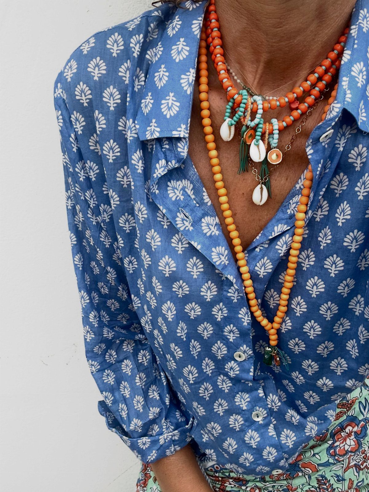 Layered orange beaded necklaces with cowrie shells worn over a blue patterned shirt.