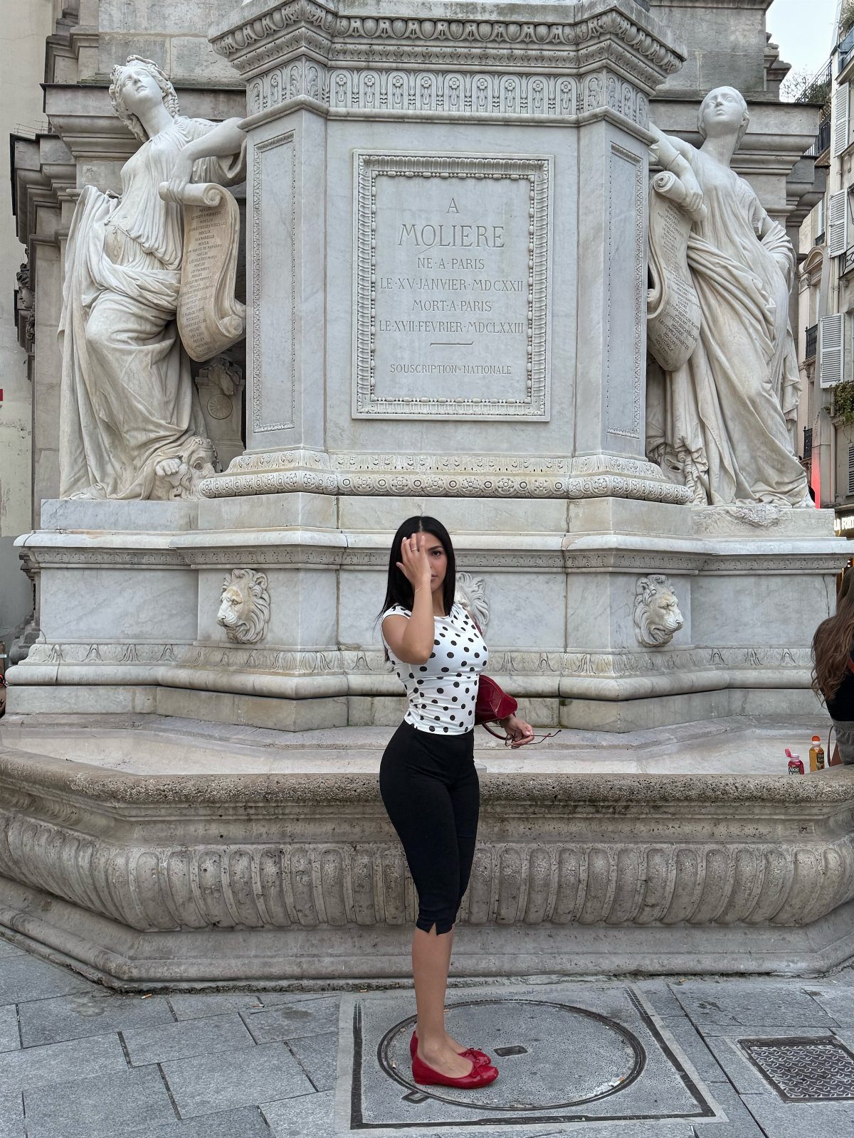 A woman wearing black capri pants and red flats poses in front of a stone monument.