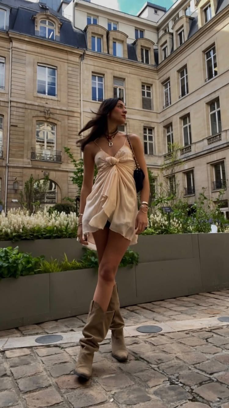 Woman in a cream draped dress and tall beige boots standing on a cobblestone street.