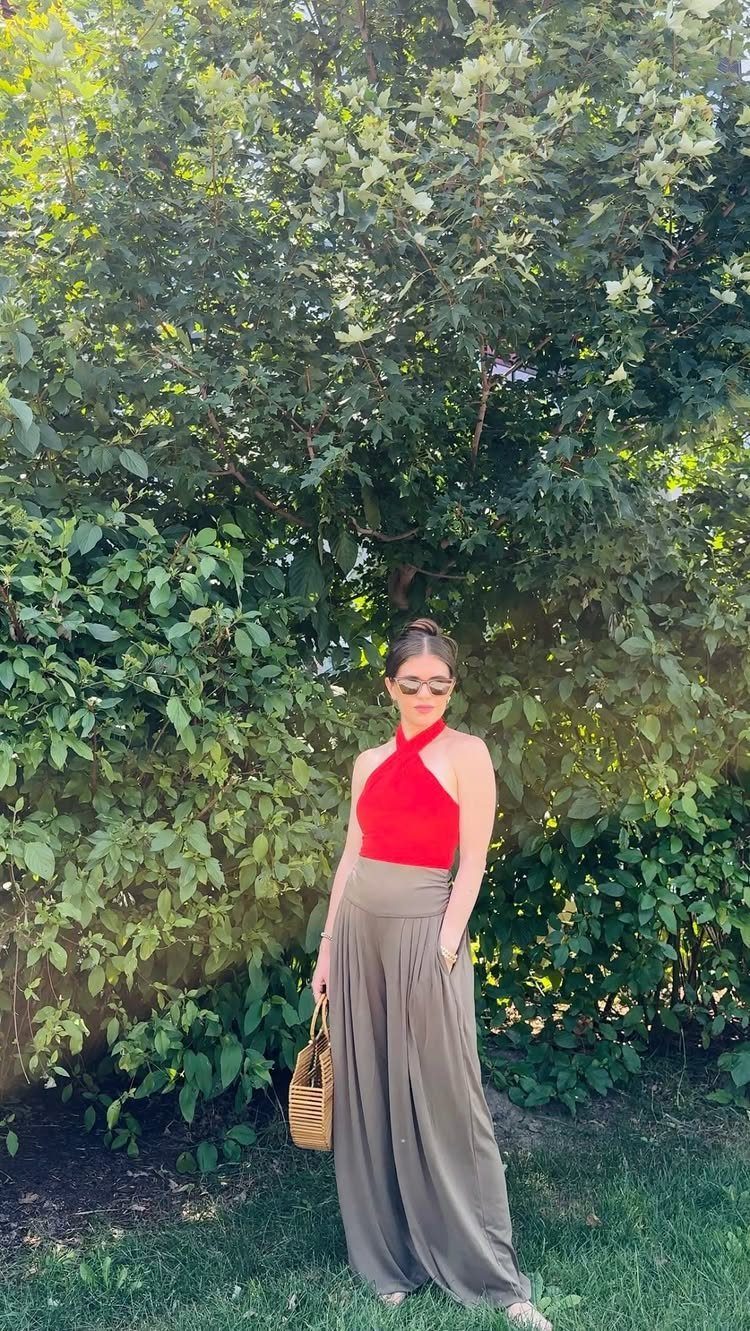 Woman in a red halter top and olive wide-leg trousers standing by green bushes.