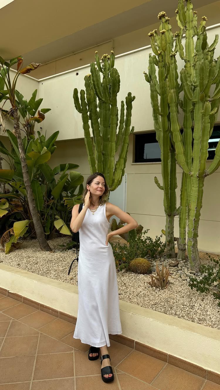 A woman in a long white linen dress standing among large cacti and tropical plants.