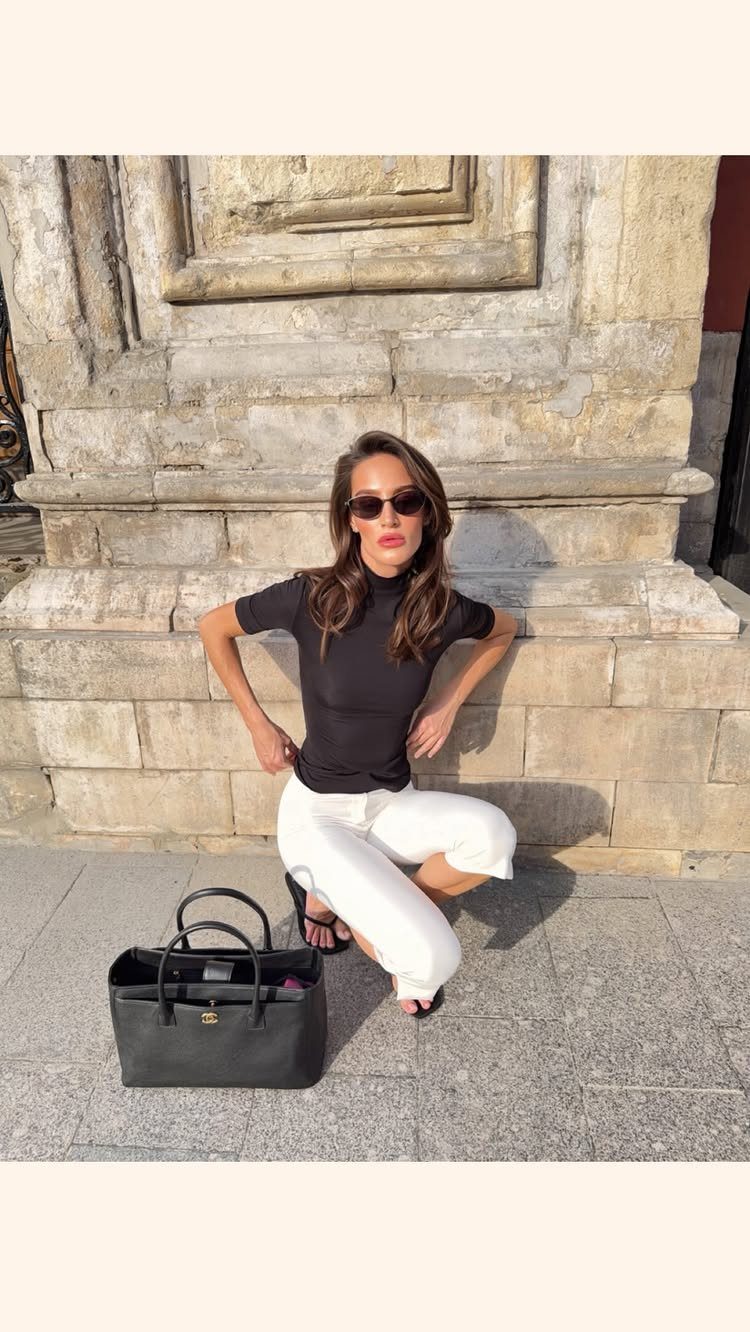Woman in black turtleneck and white trousers posing with a black handbag against a stone wall.