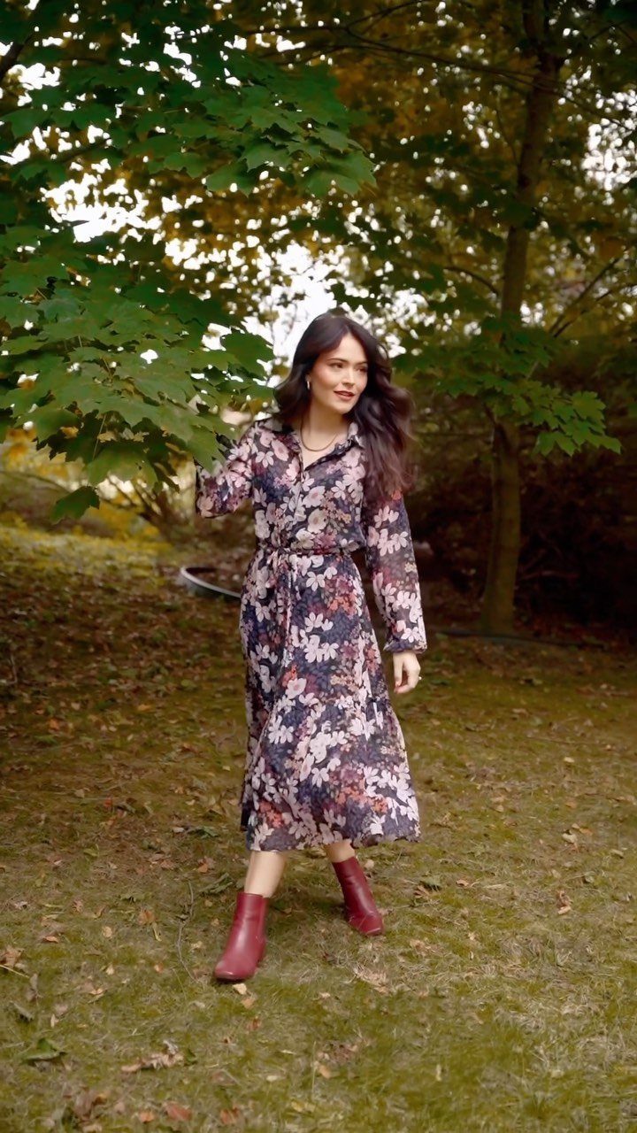 A woman wearing a floral chiffon maxi dress and red boots standing in a park.