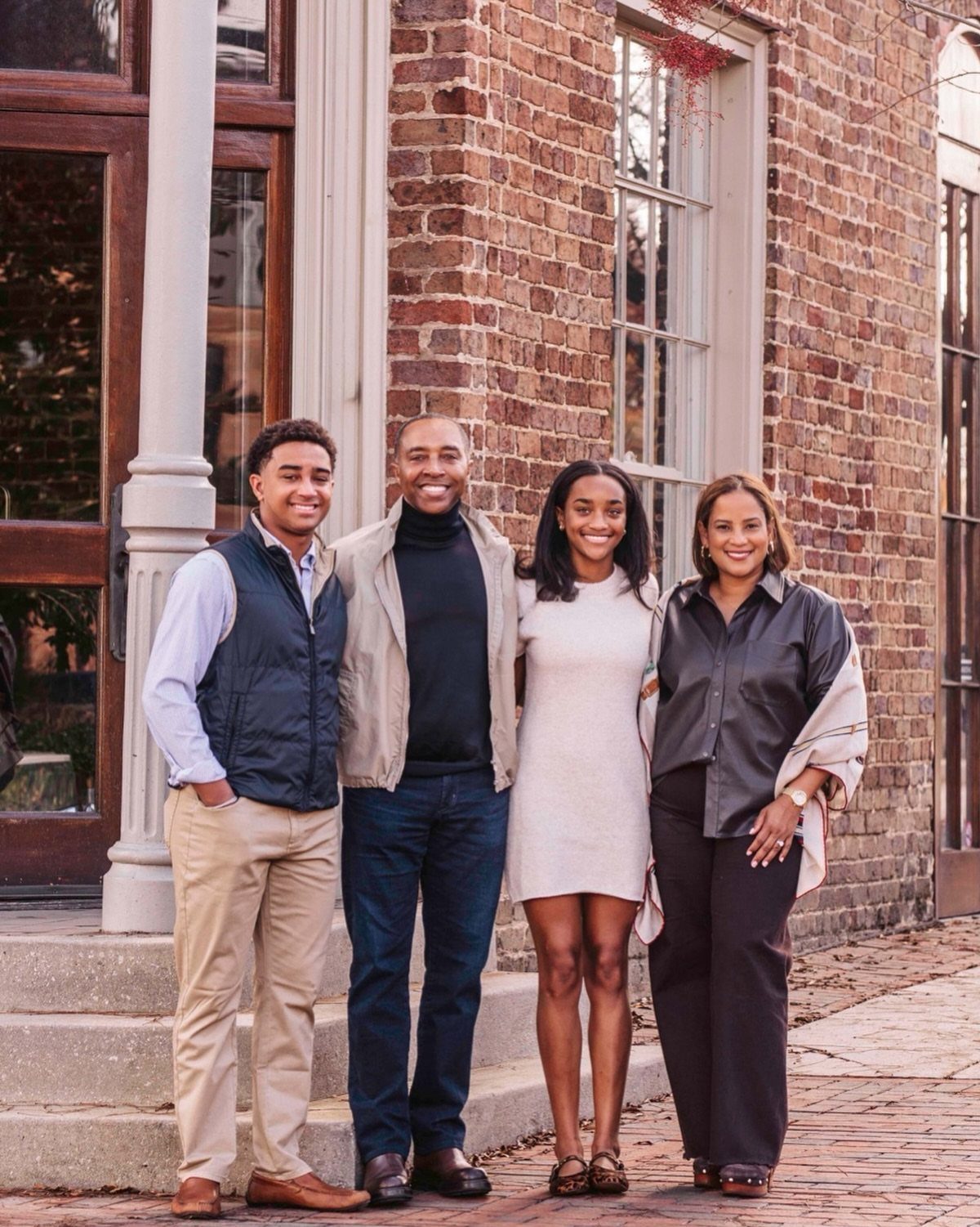 A smiling family of four posing together in front of a brick building wearing fall clothing.
