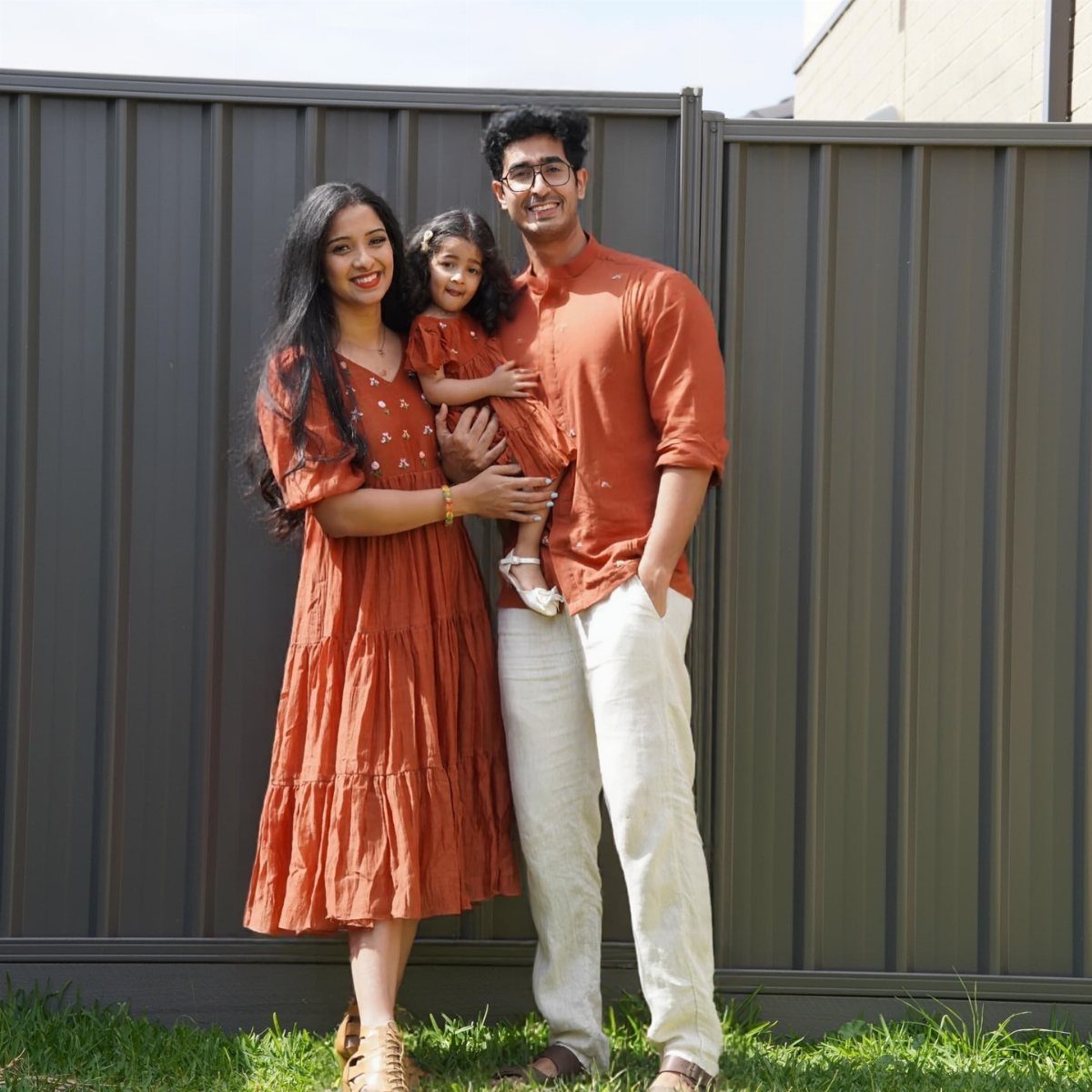 A smiling family of three wearing coordinated rust and cream autumn outfits outdoors.