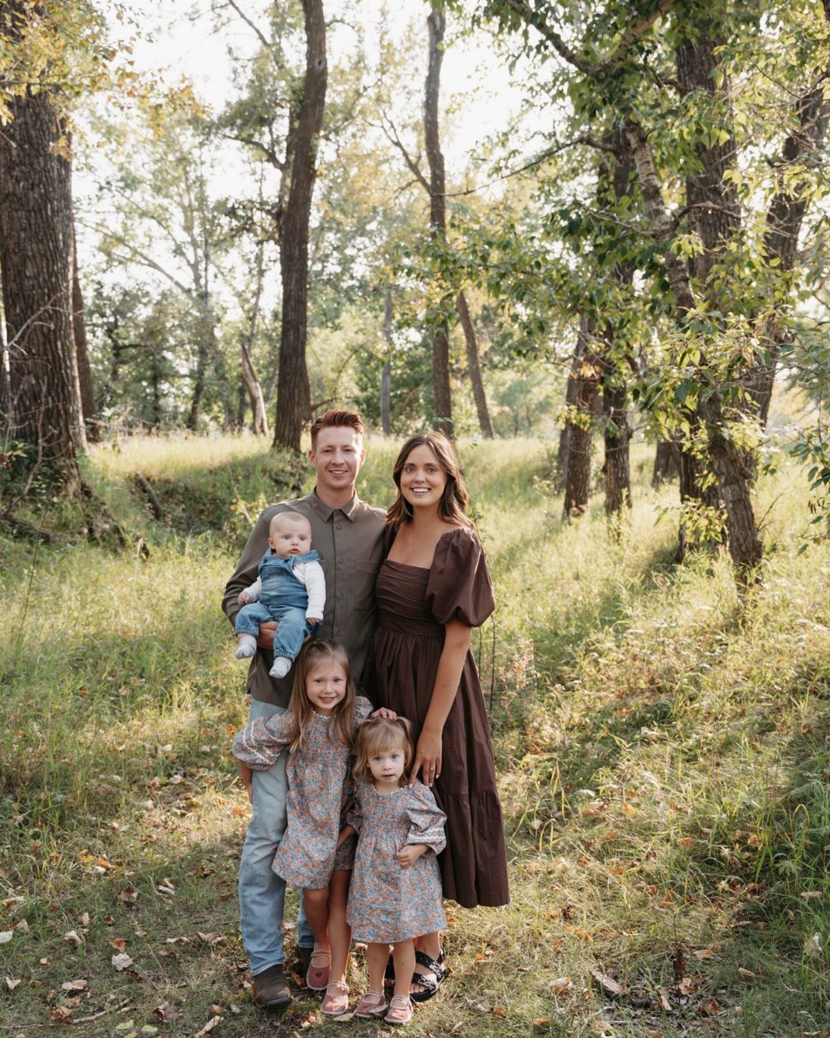 A family of five posing together in a sunlit wooded park wearing earthy toned clothing.