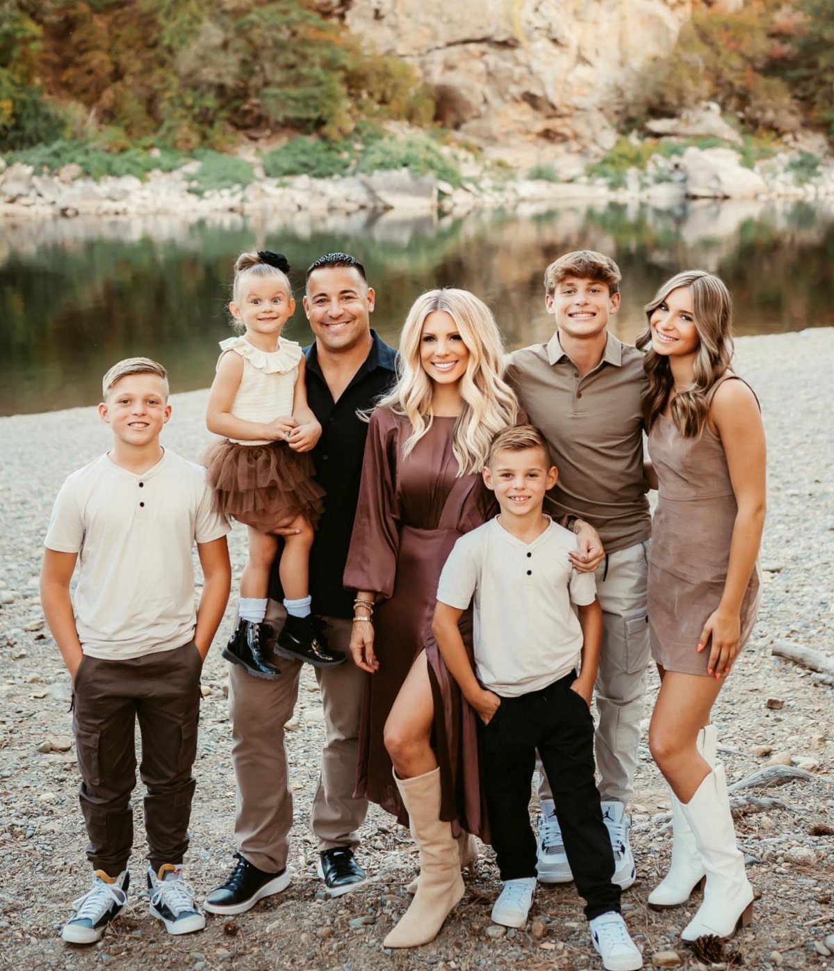 A family of seven posing together in coordinated earth-toned outfits by a river.