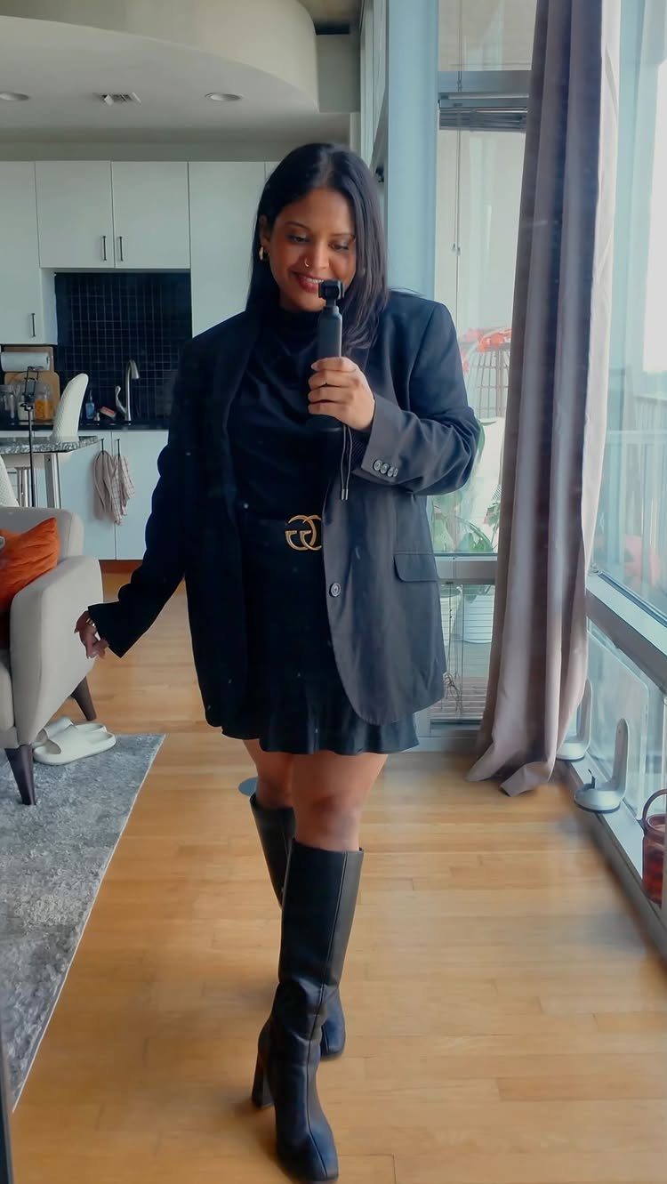 A woman posing in a black blazer, mini skirt, and tall black boots indoors.