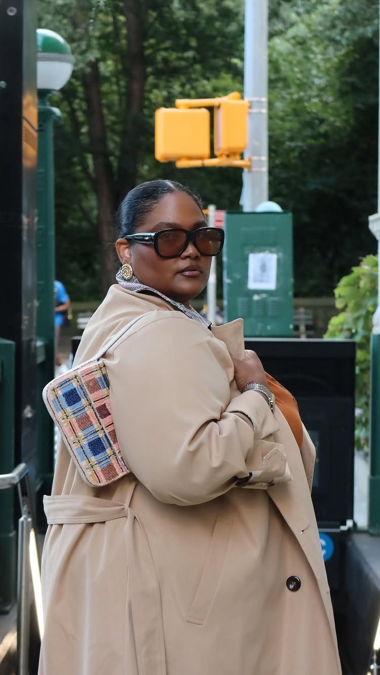 A plus-size woman wearing a tan trench coat and sunglasses posing outdoors.