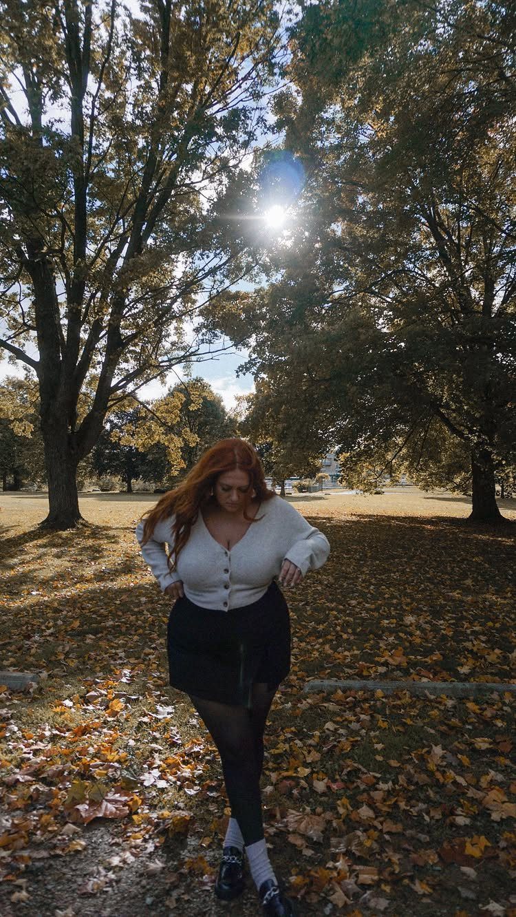 A woman with red hair posing in a park filled with autumn leaves and tall trees.