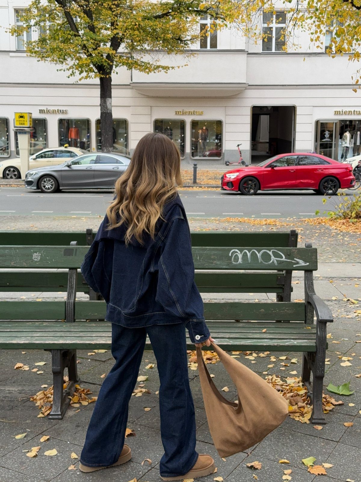 Woman wearing dark-wash wide-leg jeans and a denim jacket standing by a green bench.
