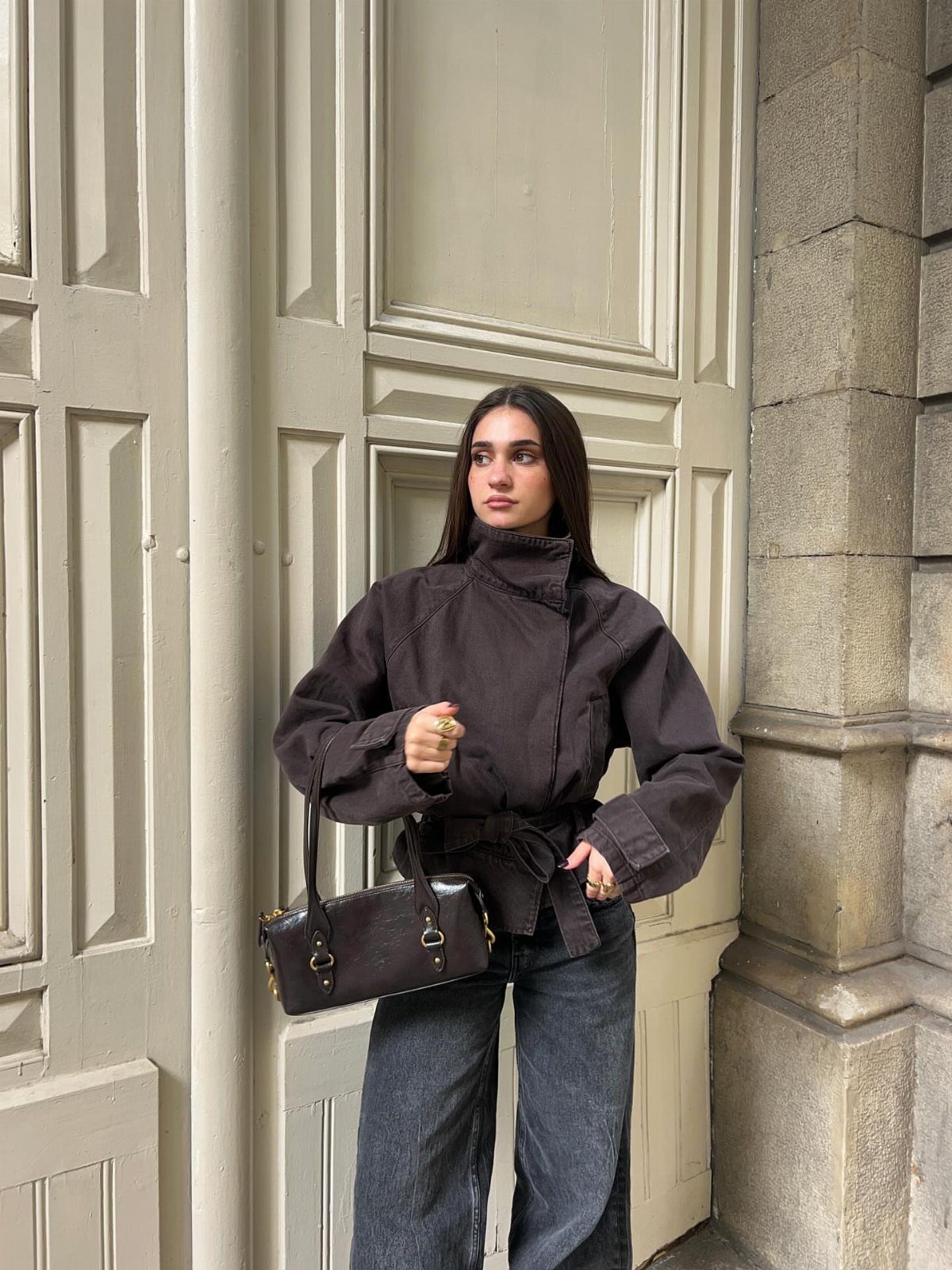A woman wearing a dark brown funnel-neck jacket and wide-leg jeans standing by a door.