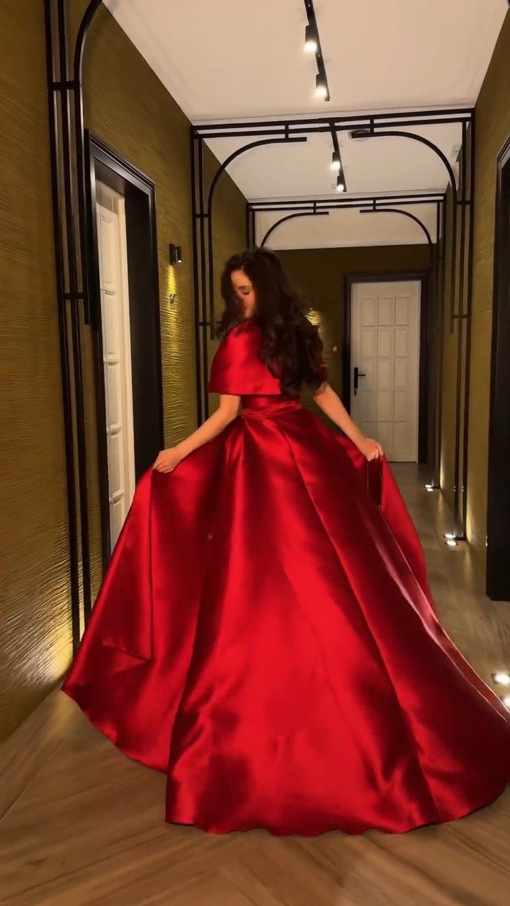 A woman in a large, flowing red satin gown standing in a modern hallway.
