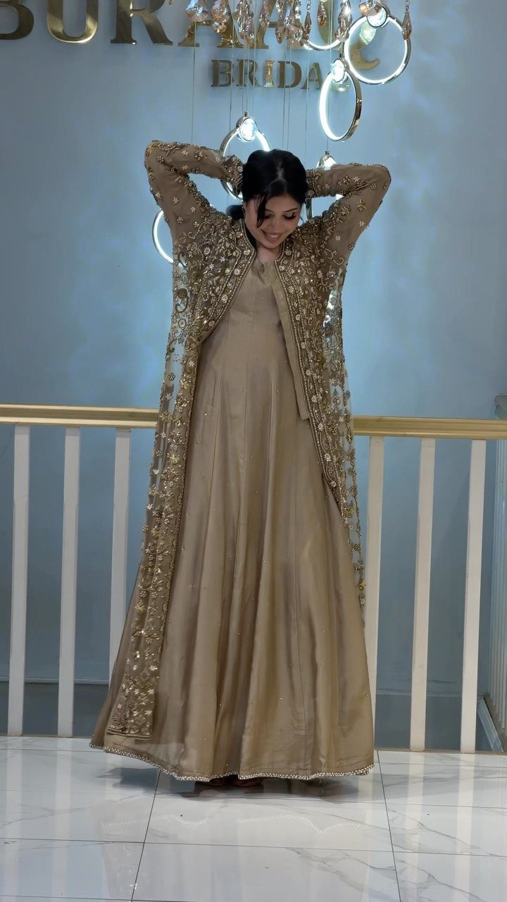Woman in an embellished gold gown and long embroidered jacket standing indoors.