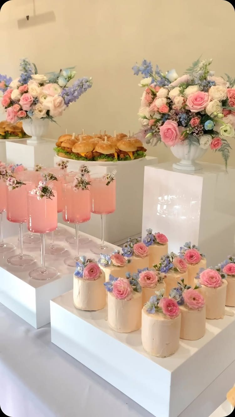 A catered party spread with pink floral cocktails, mini cakes with roses, and sliders.