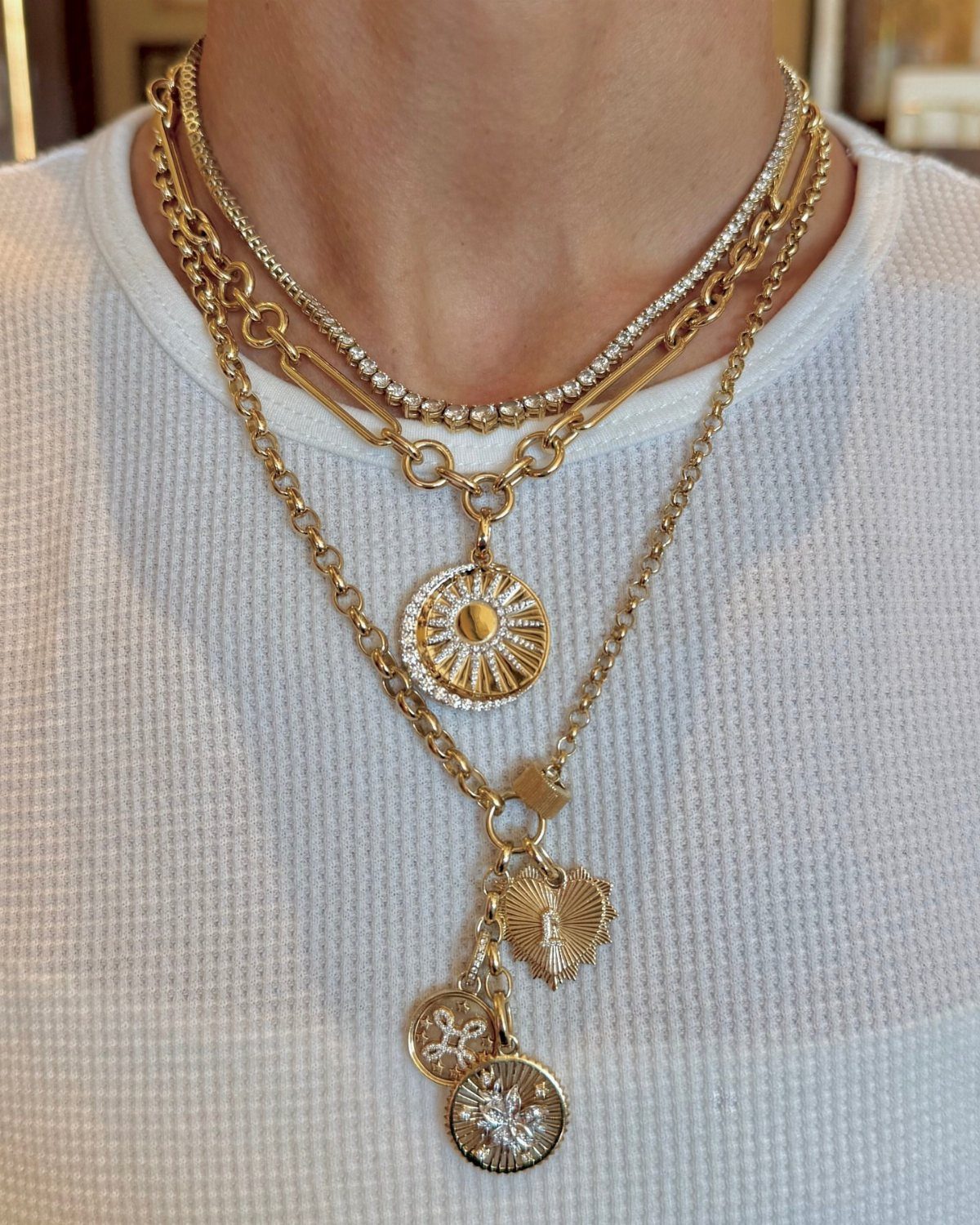 Multiple gold layered necklaces with sunburst and heart pendants worn over a white top.
