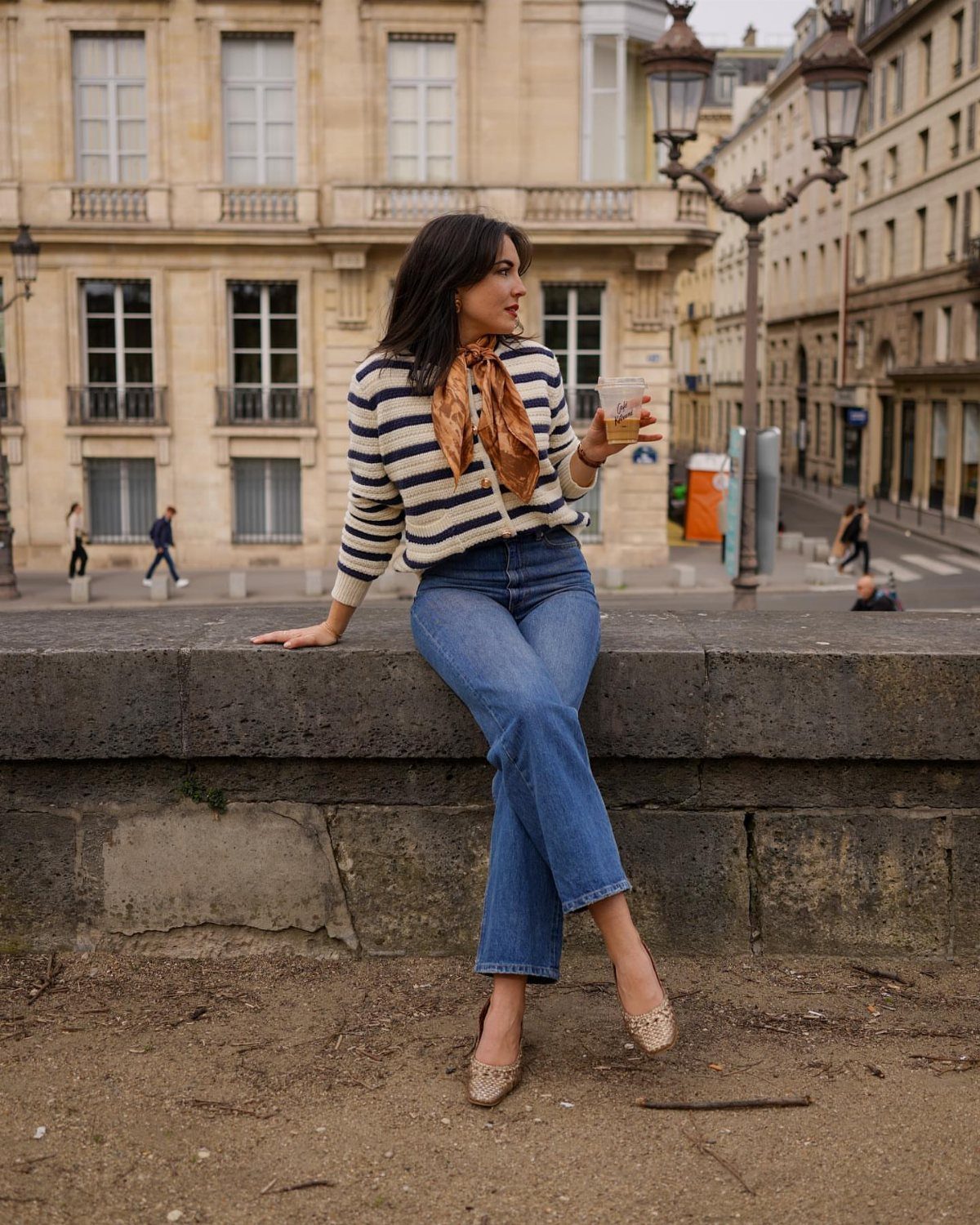 Woman wearing a striped sweater tucked into high-waisted blue jeans sitting on a stone ledge.