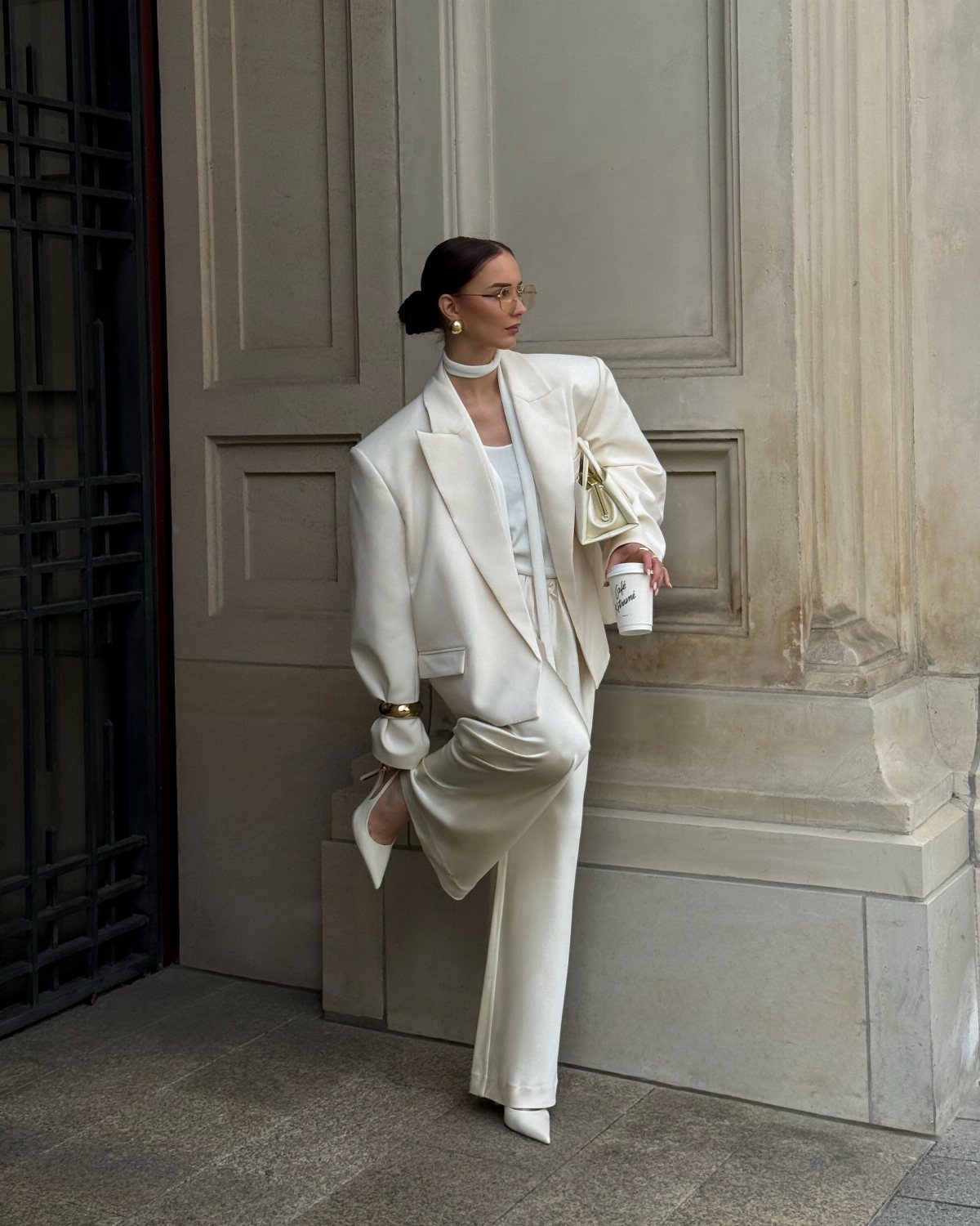 Woman in an oversized white blazer and matching wide-leg trousers leaning against a stone wall.