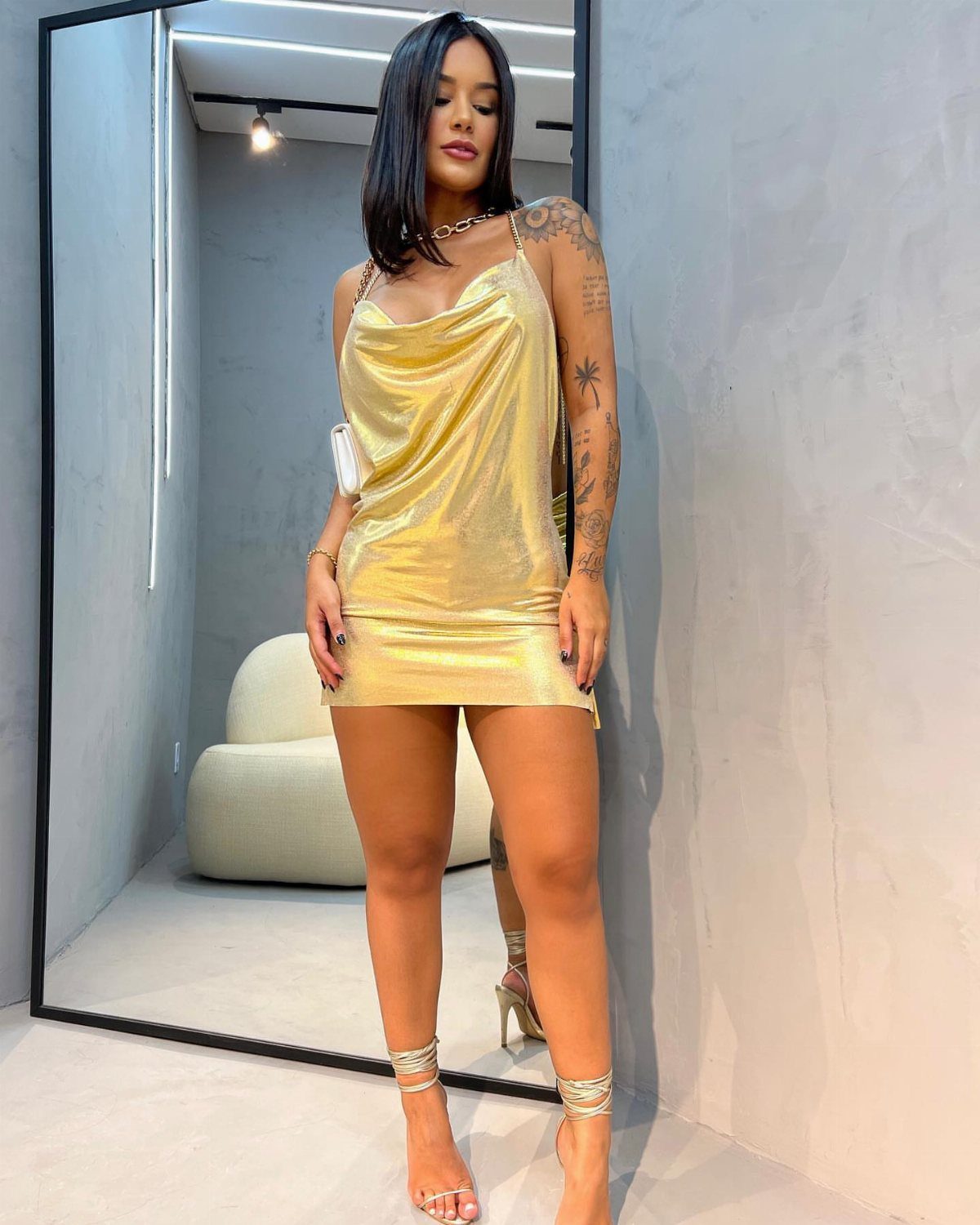 A woman posing in a short metallic gold satin dress and strappy gold high heels.