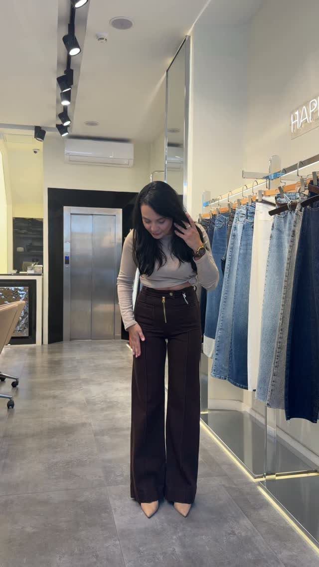 A woman wearing dark brown high-waisted wide-leg trousers and a beige long-sleeved top in a boutique.