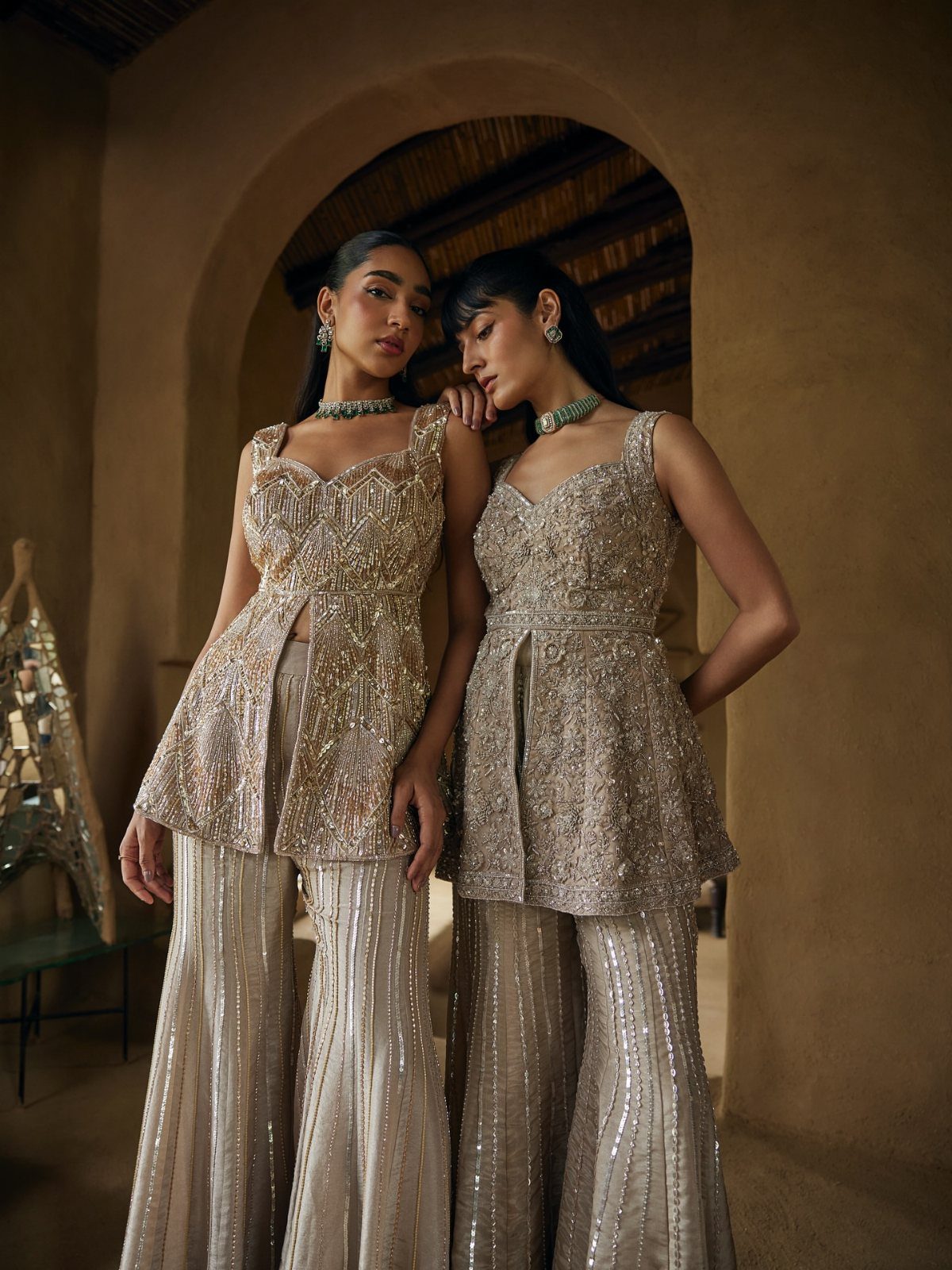 Two women posing in champagne sequined peplum tops and matching flared trousers against an archway.