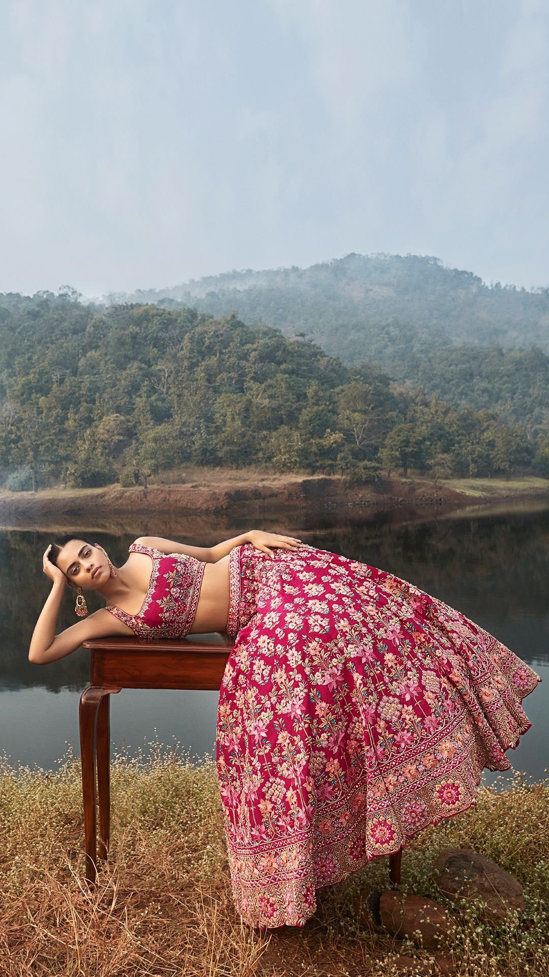 Woman in a red floral print silk lehenga lying on a wooden table by a lake.