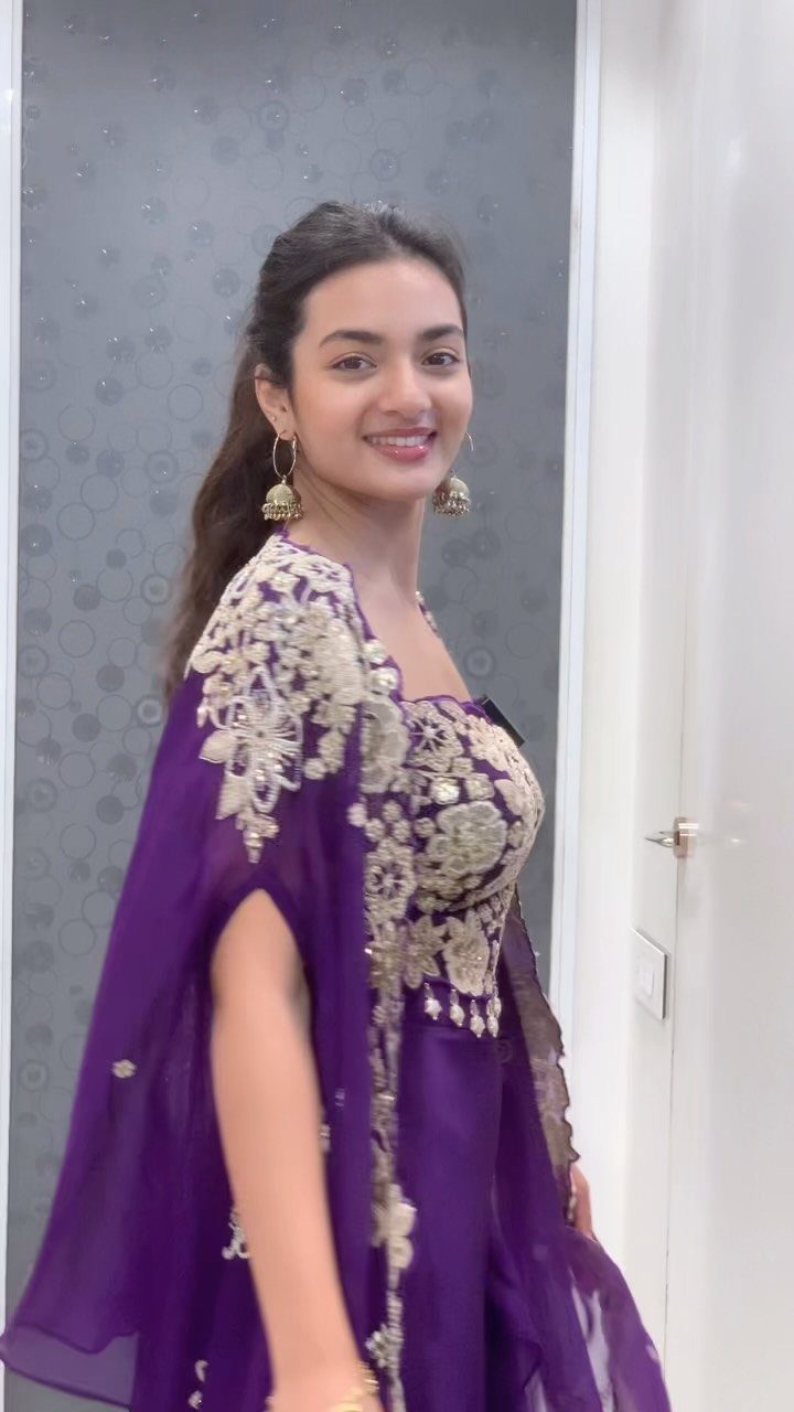 A smiling woman wearing a purple embroidered outfit with a flowing sheer cape.