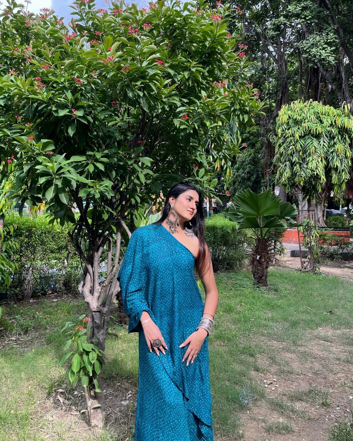 A woman in a teal one-shoulder ethnic dress posing next to a green tree.