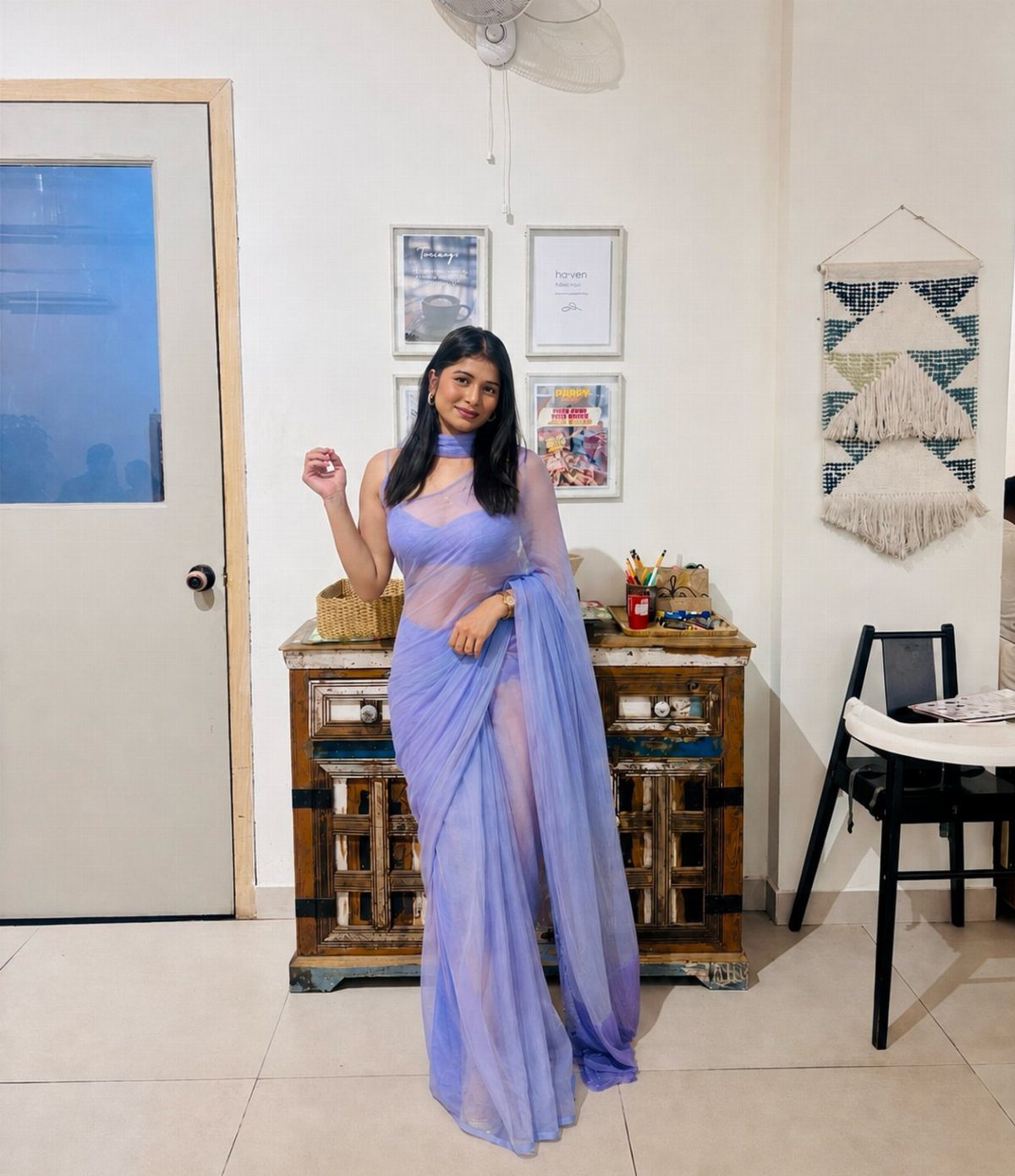 A woman wearing a flowing lavender chiffon saree posing in a decorated room.