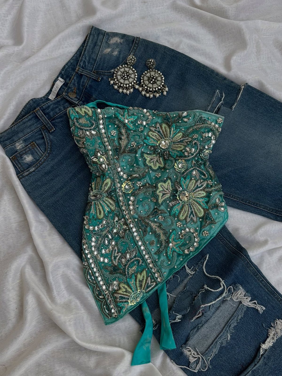 Embroidered teal corset top with silver beadwork resting on blue distressed denim jeans.