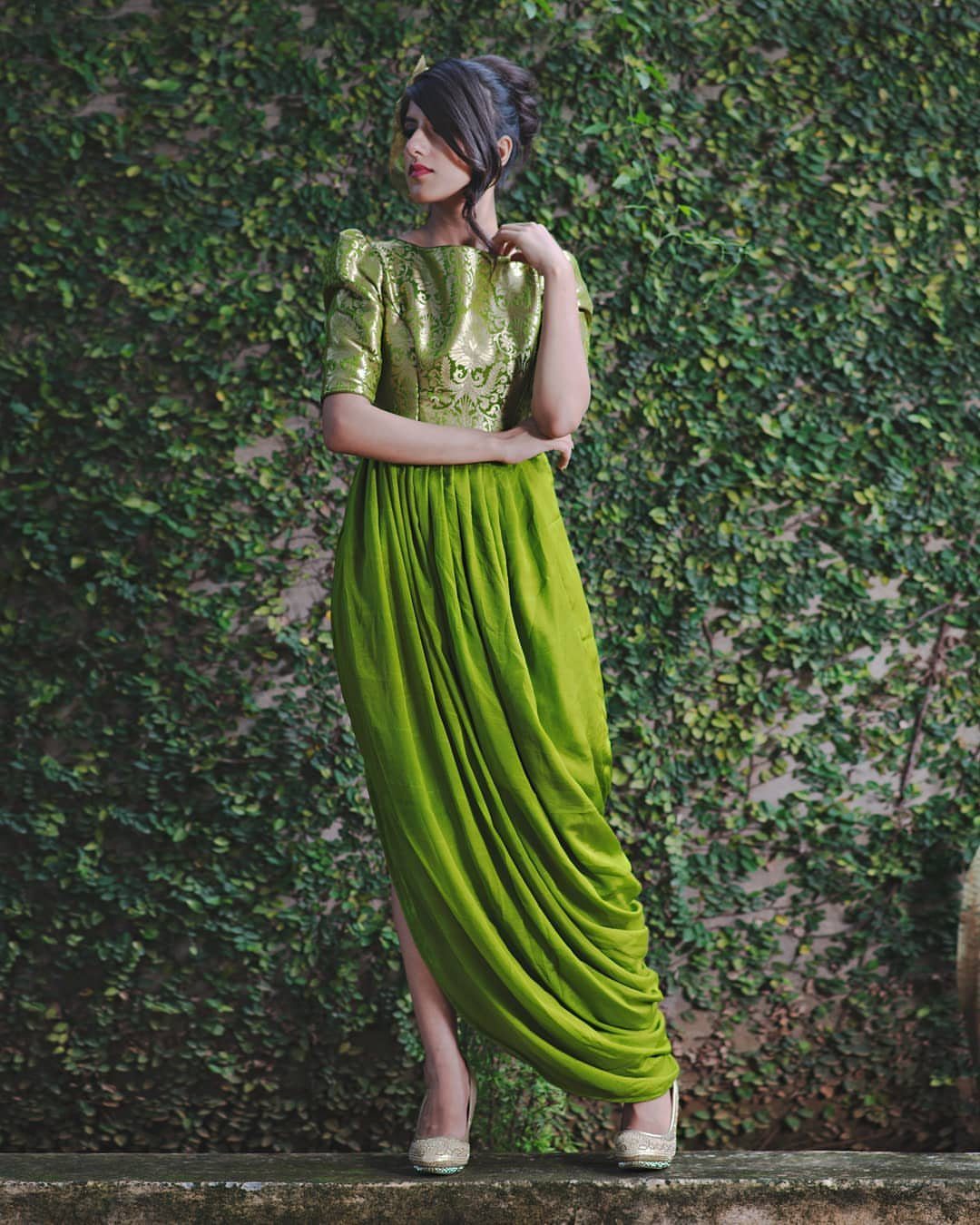 Woman in a lime green metallic top and draped dhoti pants standing against an ivy wall.