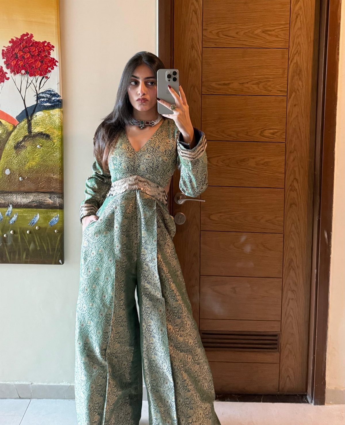 Woman in a green patterned jumpsuit posing for a mirror selfie indoors.