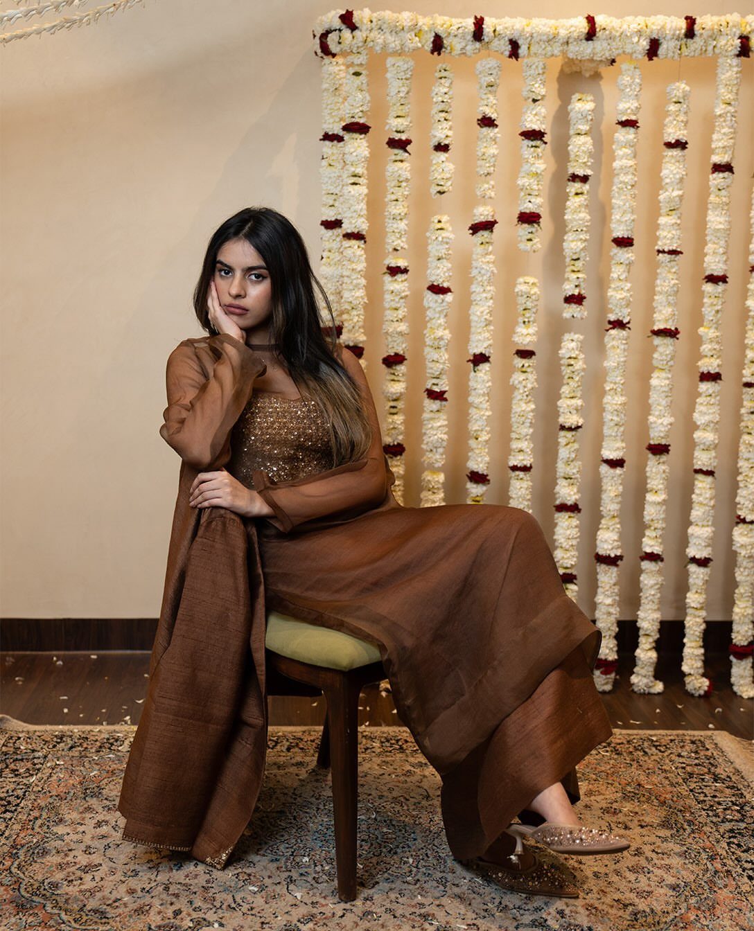 Woman in a brown sequined top and flowing skirt sitting on a chair.