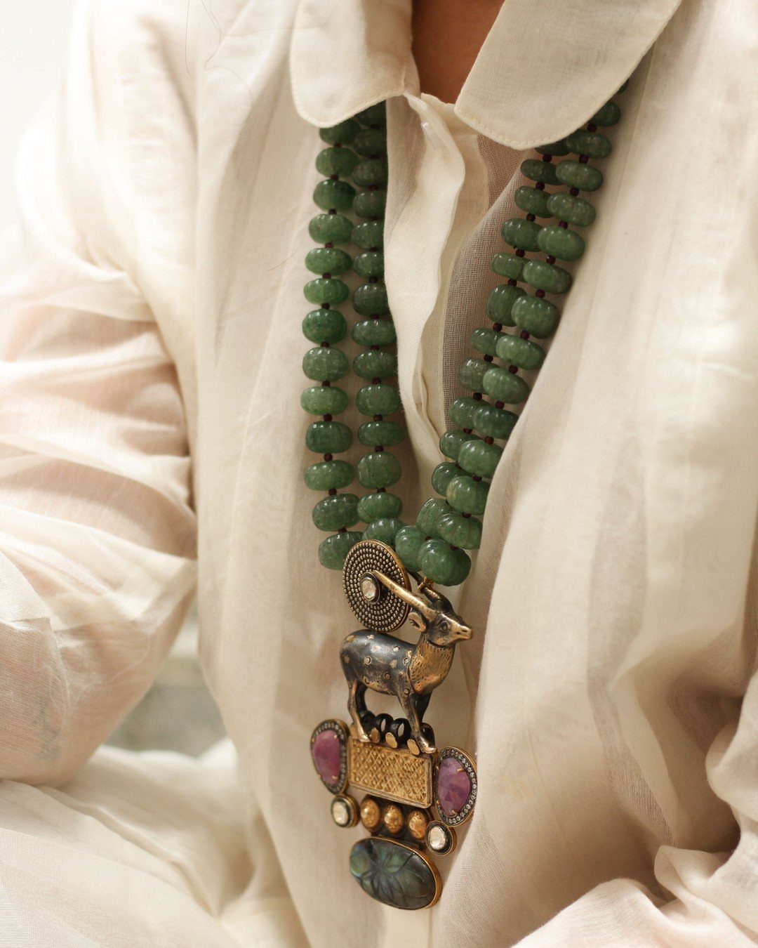 A large green beaded necklace with a gold ram head pendant and colorful gemstones worn over a white shirt.