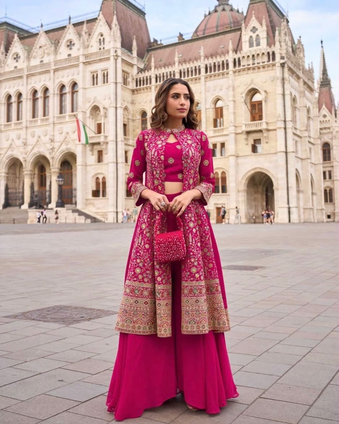 Woman wearing a pink embroidered three-piece Indo-Western outfit standing in front of a large building.