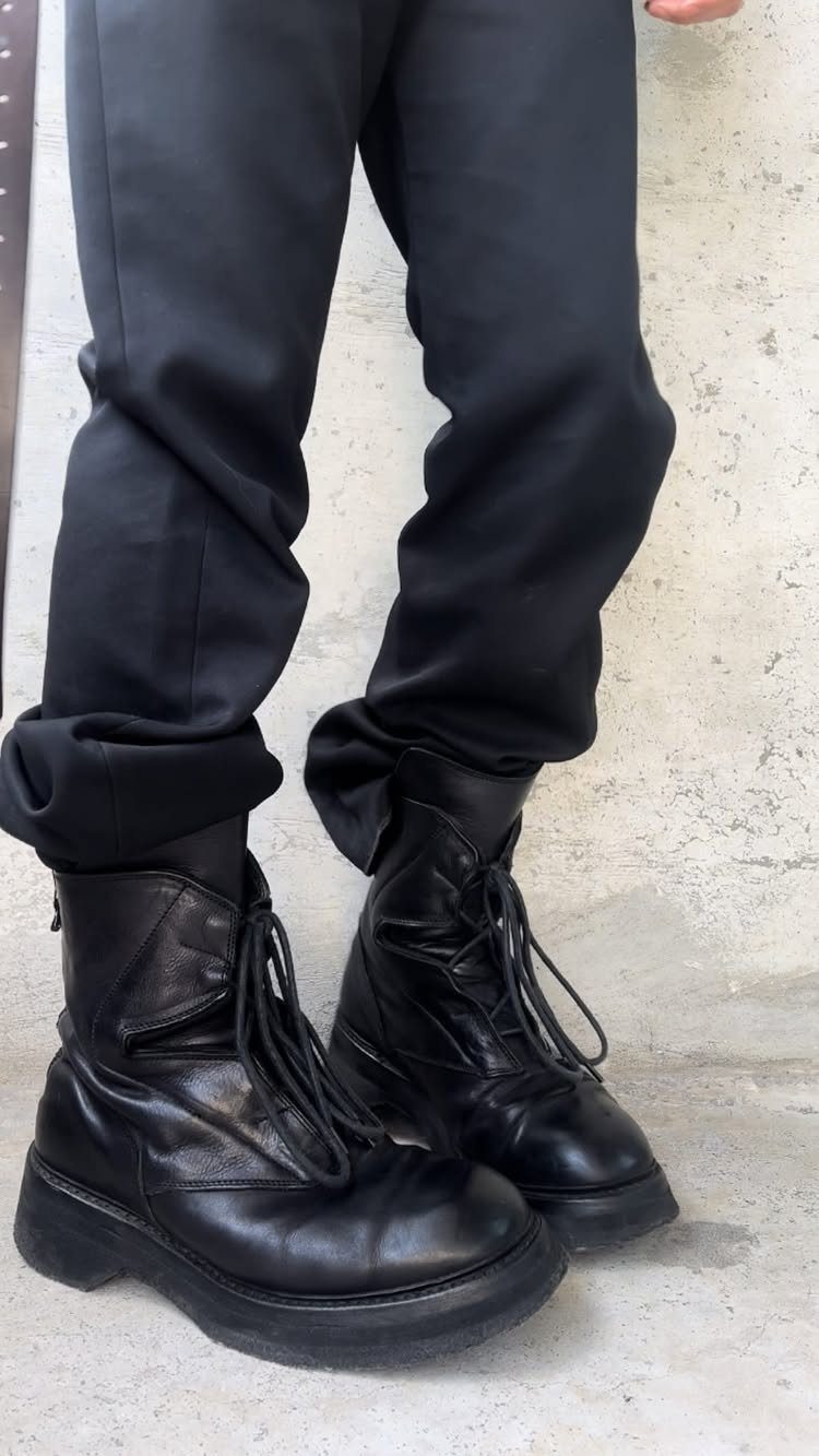 Black leather combat boots worn with black trousers against a textured wall.