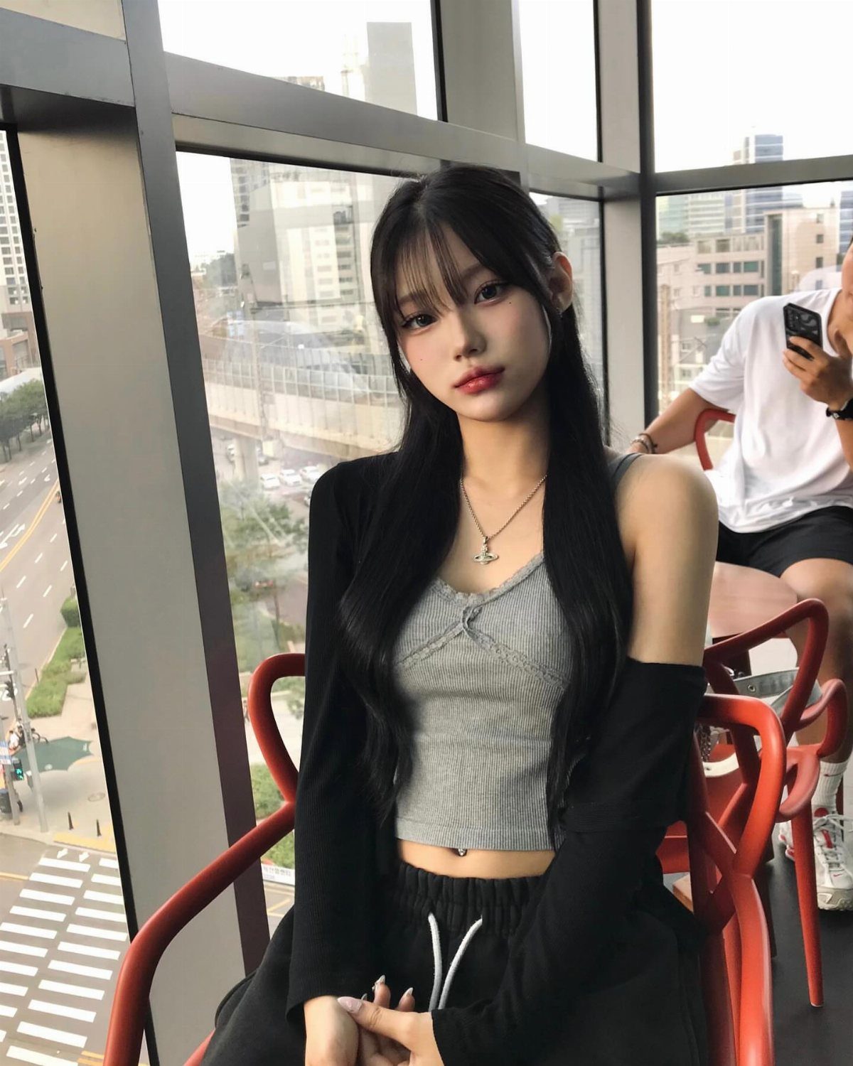 A young woman wearing a grey ribbed cropped tank top and black cardigan sitting by a window.