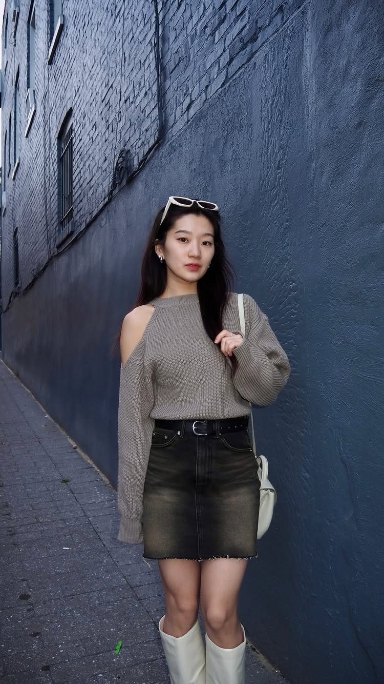 A woman wearing a beige sweater and a dark denim mini skirt standing against a blue wall.