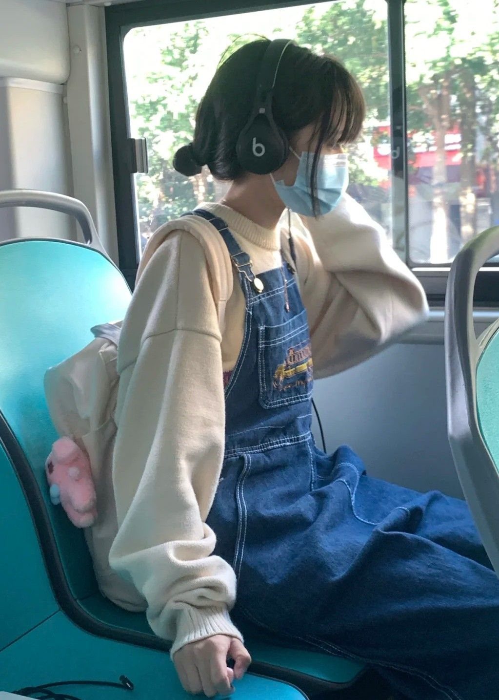 Person in cream oversized sweater and denim overalls sitting on a bus wearing headphones.