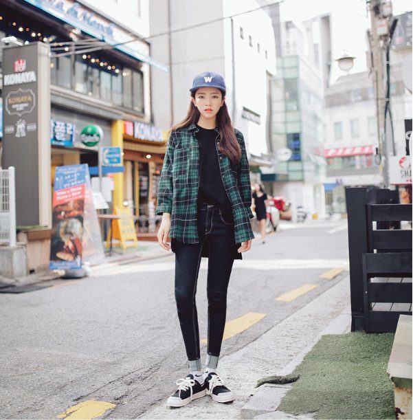 A woman wearing a green plaid shirt, black jeans, and a blue baseball cap on a city street.