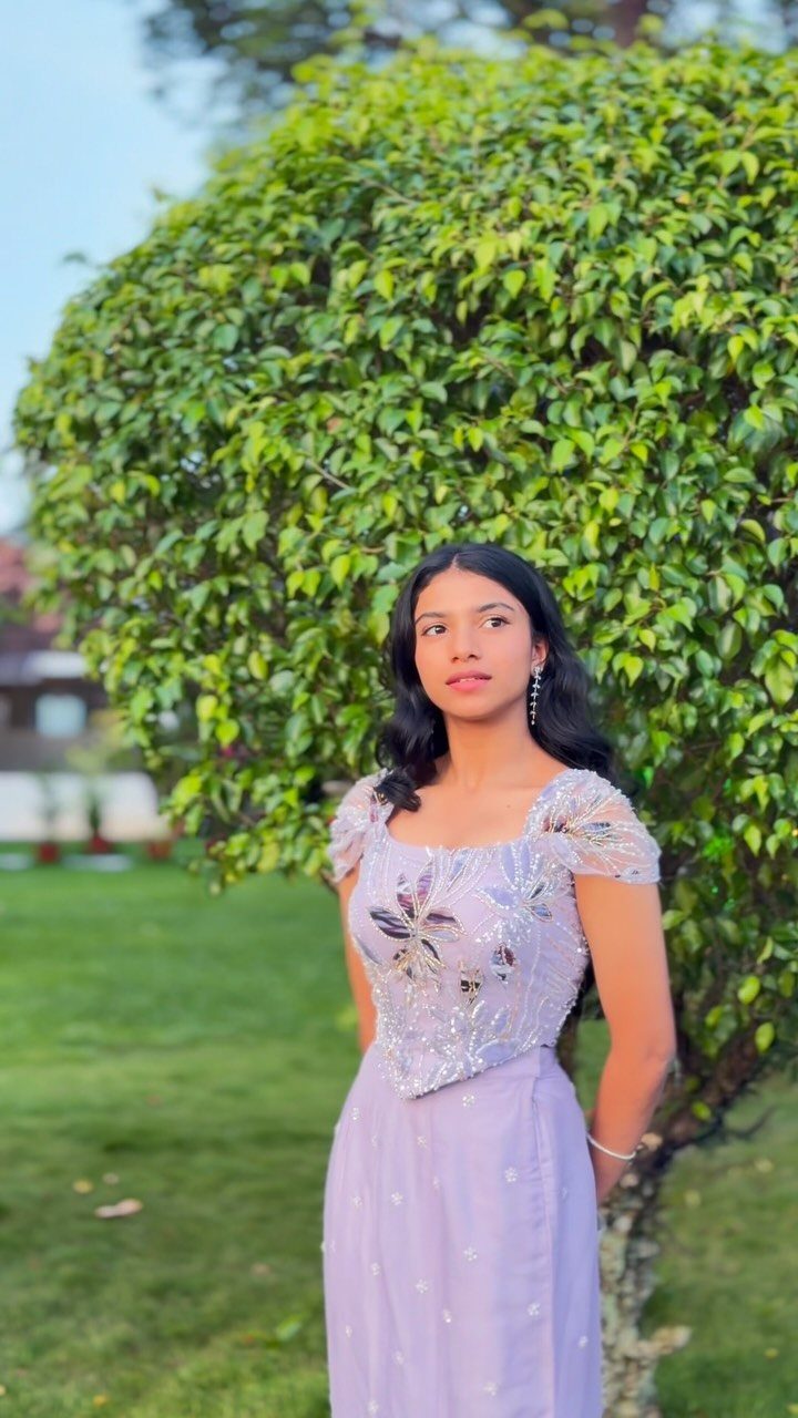 A young woman posing in a lavender dress with silver floral embroidery against a green bush.