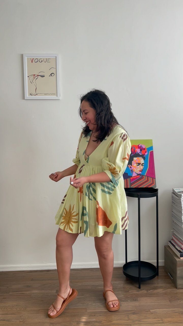 A woman smiling while wearing a short, patterned, light yellow babydoll dress and brown sandals.
