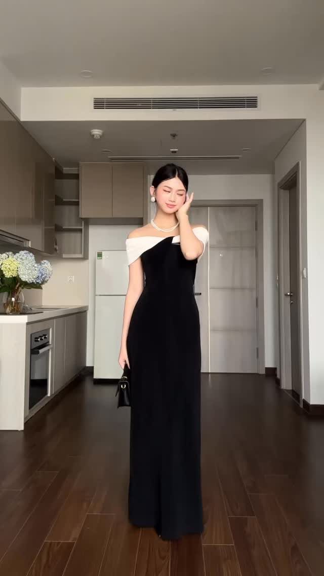 A woman in a black and white off-the-shoulder maxi dress standing in a modern apartment.